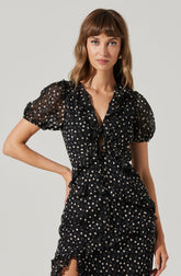 A woman is wearing a black dress with short, puffed sleeves made of sheer fabric. The dress features a polka dot pattern with small, light-colored dots scattered throughout. It has a ruffled design with layered fabric and a front drawstring detail at the neckline.